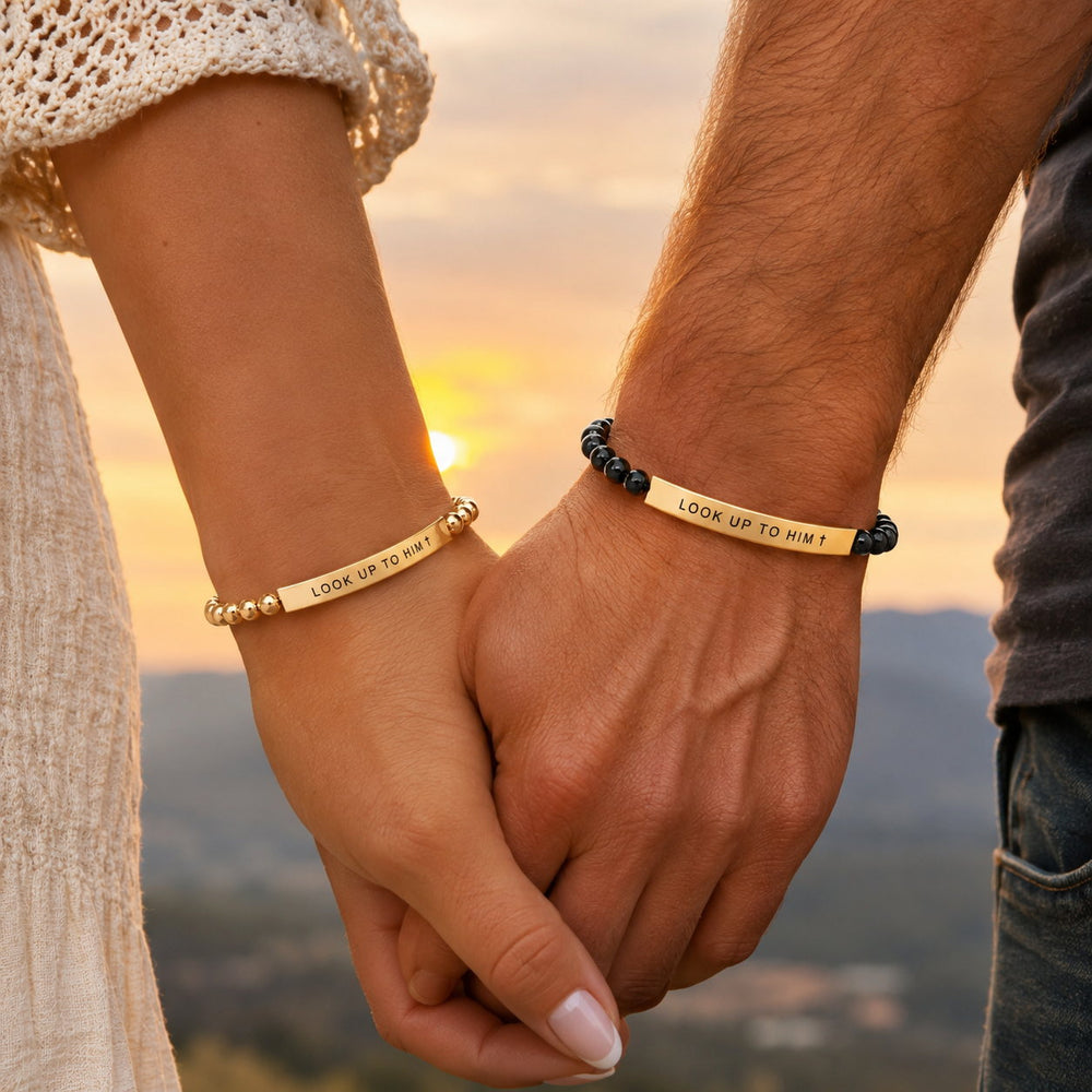 Couple holding hands at sunset wearing matching Look Up to Him bracelets, Gold for her and Black & Gold for him